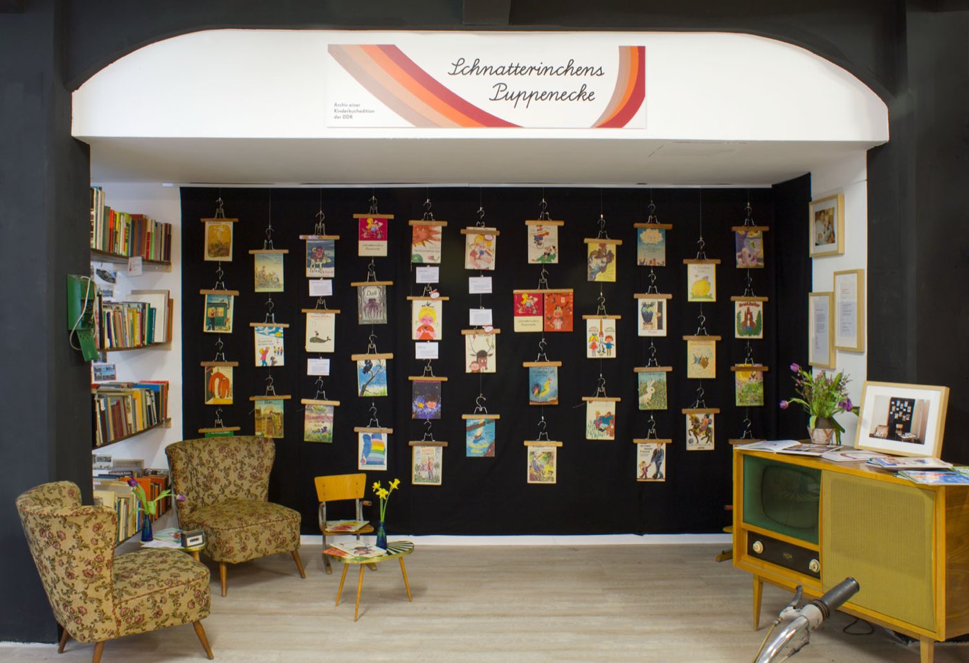 Ausstellungsansicht im N&rsquo;OSTALGIE-Museum: an einer schwarzen Wand h&auml;ngen rund vierzig Kinderb&uuml;cher, davor Sessel und ein Radioschrank