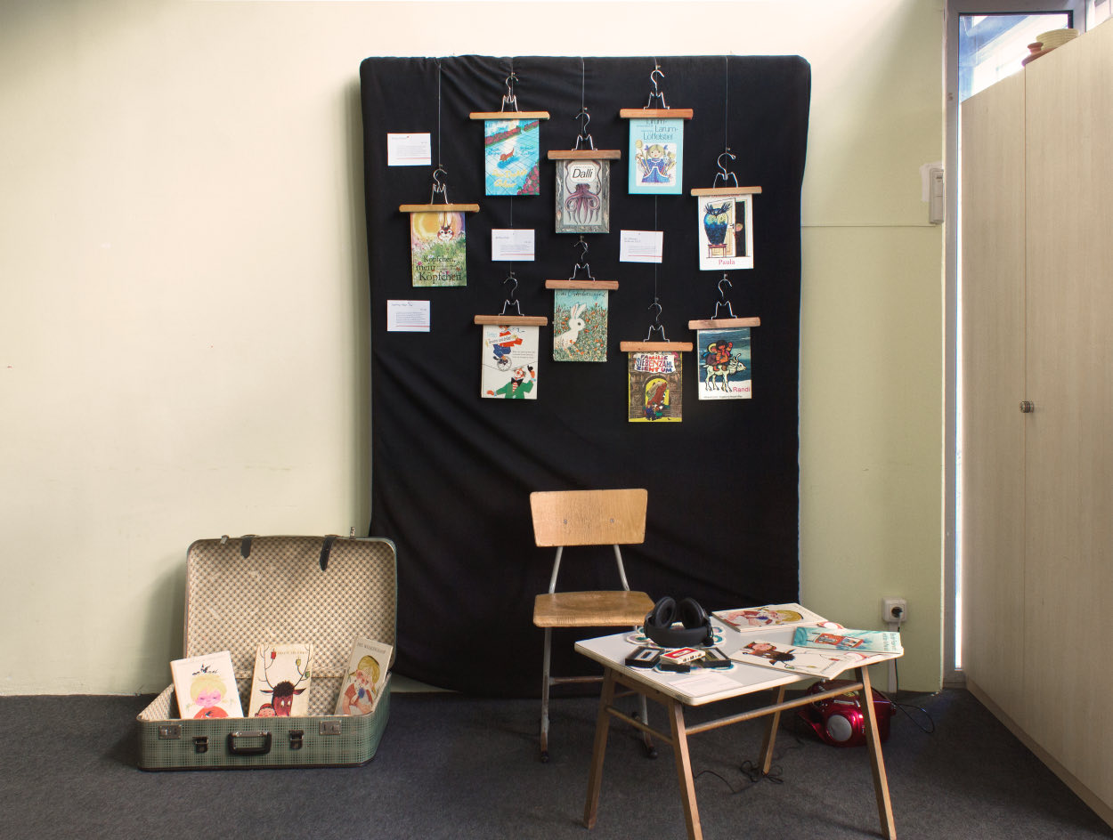 Ausstellungsaufbau: Kinderb&uuml;cher an einer schwarzen Stoffwand, davor ein Koffer, ein Kinderstuhl und ein Tisch